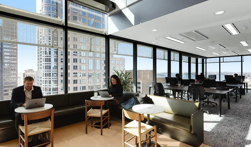 People work on laptops at small round tables along a banquette inside a modern high-rise office with large floor-to-ceiling windows overlooking city skyscrapers and a harbor.