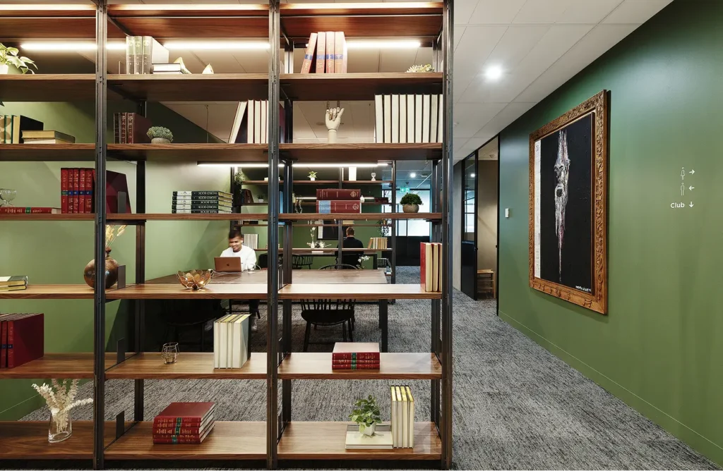 A wooden open-shelved bookcase acts as a room divider, separating a carpeted office hallway from a seating area where individuals work at tables beside green walls and framed artwork. Text on the right wall reads: "Club ↓". Text on book spines includes: "WORLD BOOK" "YEAR BOOK"