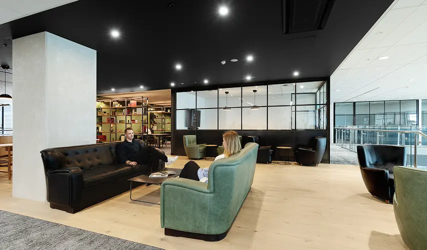 A man and a woman sit on leather sofas facing each other in a modern office lounge area featuring light wood floors, dark ceilings, and glass-partitioned rooms.