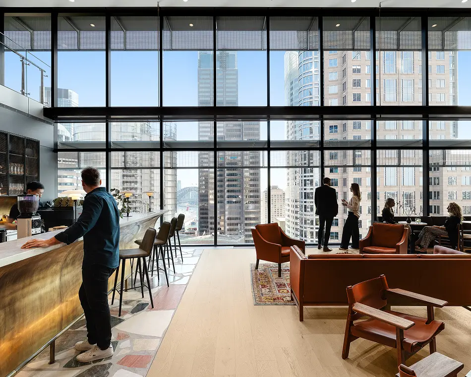 People converse and relax in a modern high-rise lounge featuring leather armchairs and a coffee bar, set against floor-to-ceiling windows overlooking city skyscrapers.