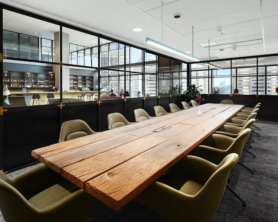 A long rustic wooden conference table surrounded by olive-green armchairs dominates a modern meeting room, enclosed by black-framed glass walls overlooking an adjacent lounge and city skyline.