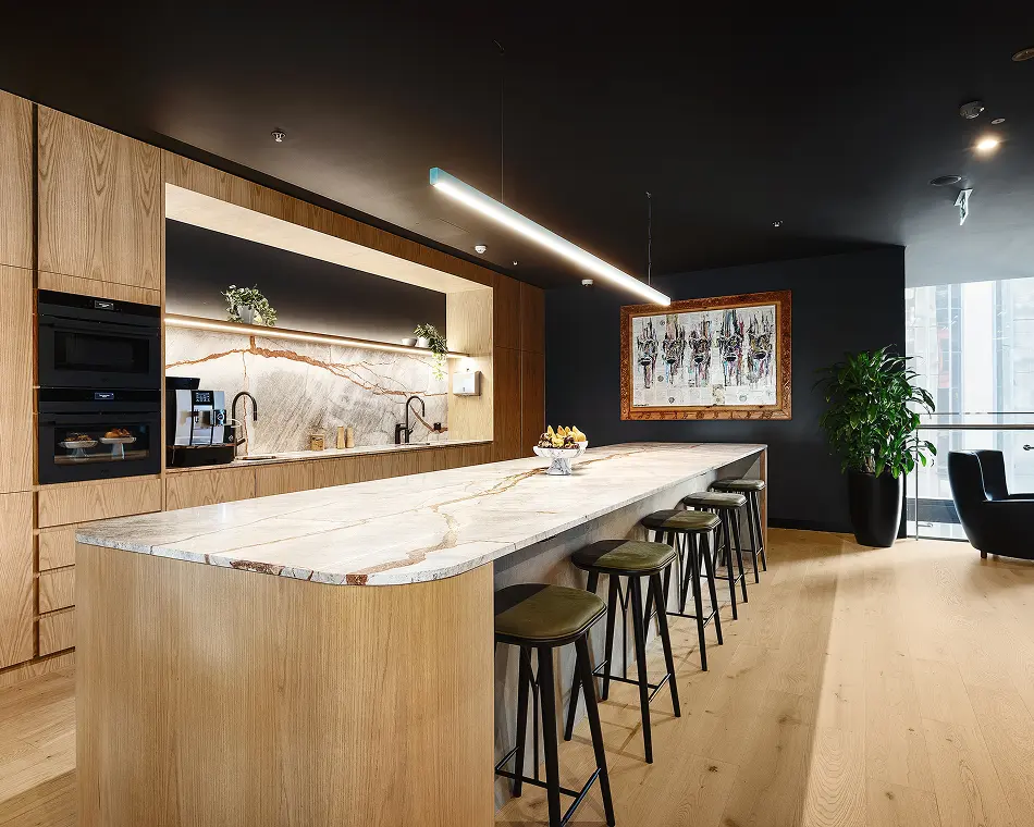 A long, marble-topped kitchen island lined with bar stools extends through a modern breakroom, framed by light wood cabinetry, built-in appliances, dark walls, and light wood flooring.