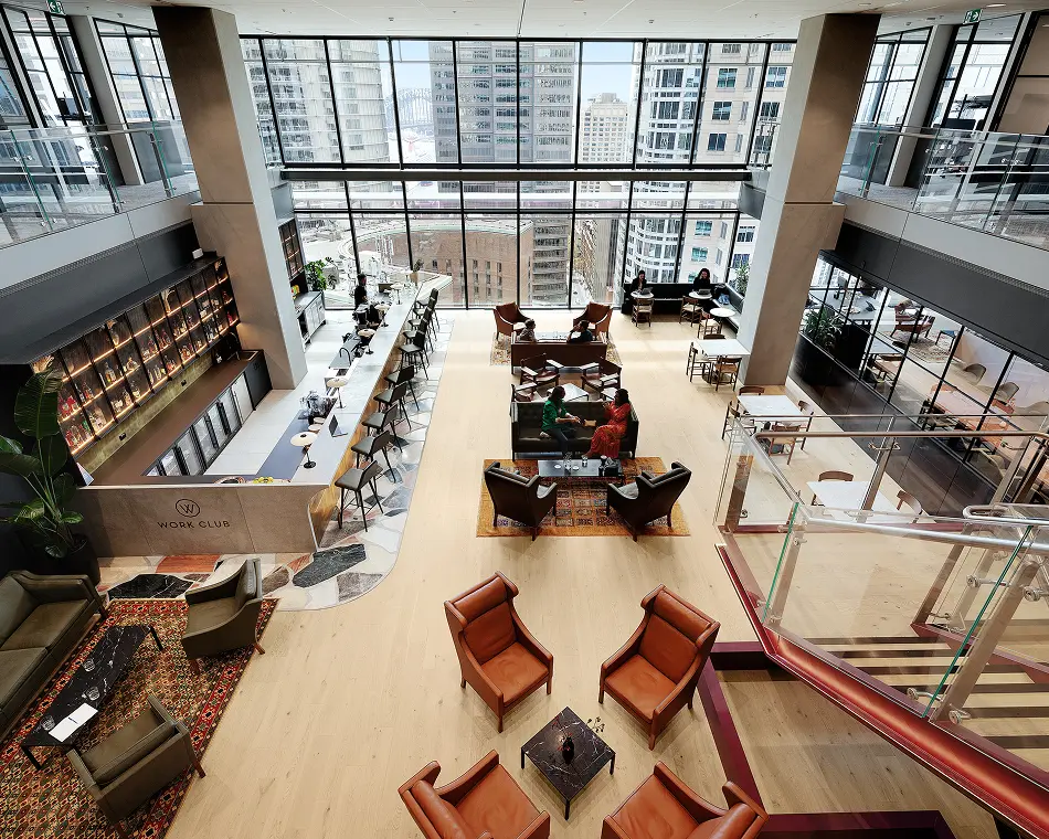 People relax and converse in a spacious high-rise lounge featuring plush armchairs, a long bar counter with text that reads "WORK CLUB" below a "W" logo, and floor-to-ceiling windows overlooking a cityscape.