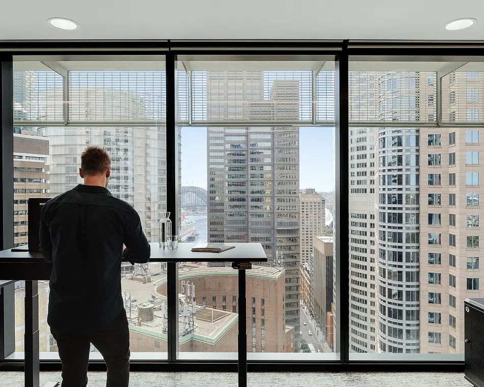 A man in dark clothing stands at a high desk, gazing out floor-to-ceiling office windows at a dense city skyline filled with tall skyscrapers.