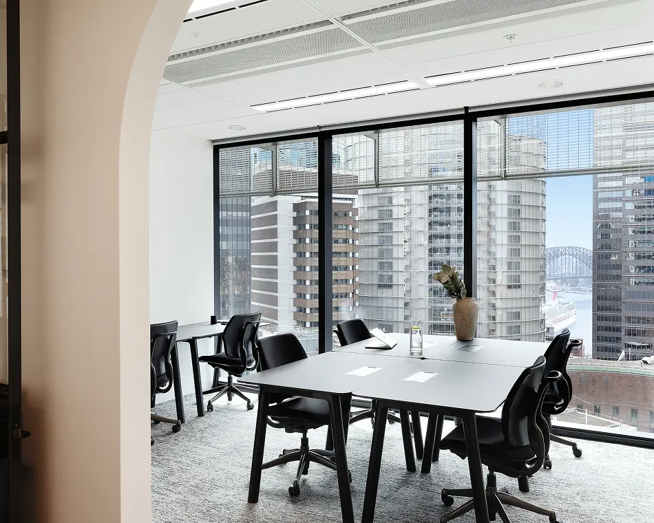 Sleek black conference tables and office chairs sit empty inside a bright workspace, framed by large windows overlooking high-rise city buildings and a distant harbor bridge.