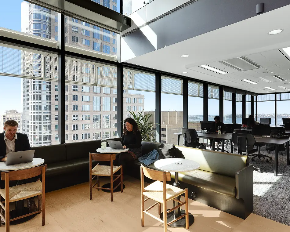 People sit at round tables along a dark green banquette while working on laptops inside a modern high-rise office with expansive floor-to-ceiling windows offering cityscape views.