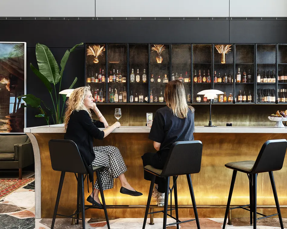 Two women sit on high black barstools, chatting over drinks at a brass-fronted bar counter inside a stylish lounge featuring an illuminated, mesh-front liquor cabinet behind them.