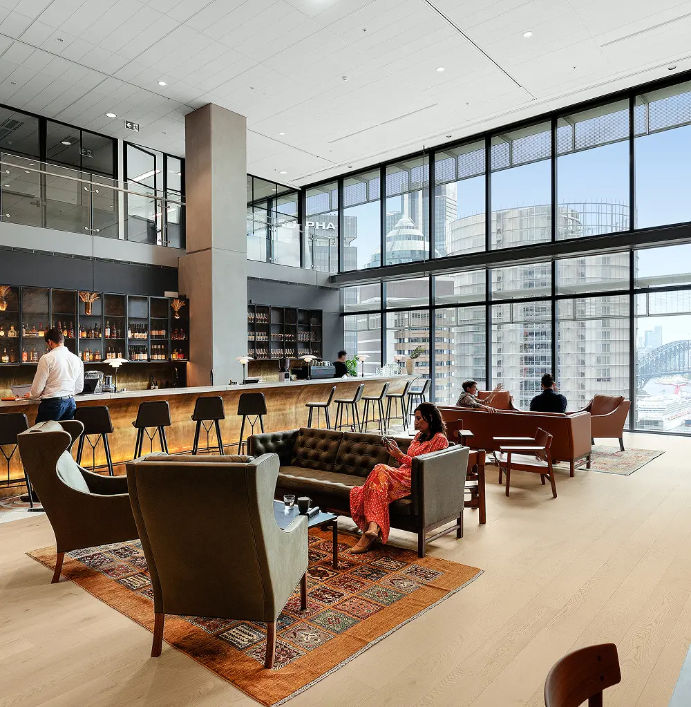 People relax in a high-ceilinged modern lounge furnished with plush sofas, armchairs, and a long bar. Floor-to-ceiling windows overlook city skyscrapers and a harbor. The text “ALPHA” is visible on an upper-level glass railing.