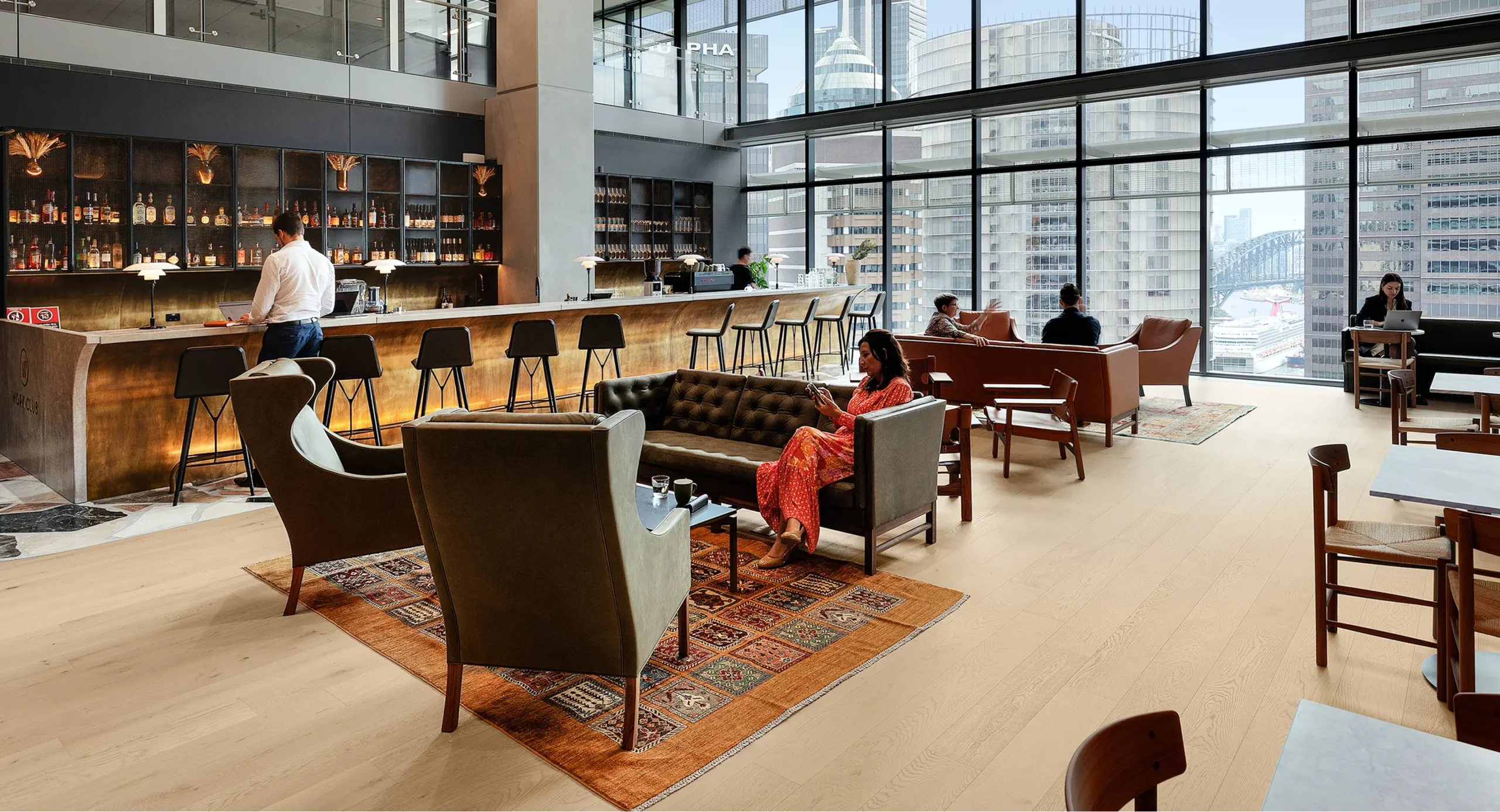 Patrons relax and work in a modern, high-rise lounge featuring a long bar, plush seating over patterned rugs, and floor-to-ceiling windows with city skyline views. Text reads "IVORY CLUB" and "ALPHA".