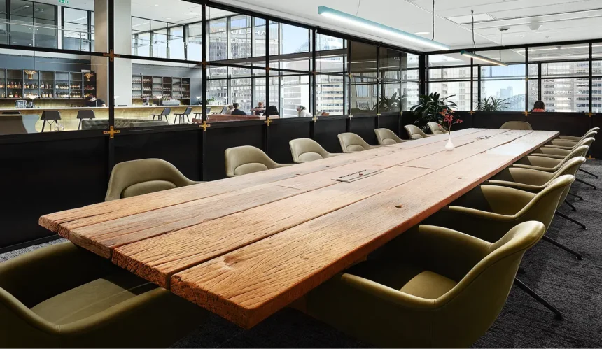 A long, rustic wooden conference table surrounded by olive-green modern armchairs stretches through a meeting room enclosed by black metal-framed glass walls overlooking an adjacent lounge and city skyline.