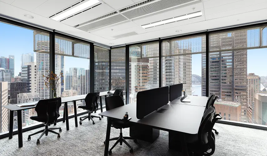 Modern black office desks and ergonomic rolling chairs sit arranged inside a high-rise office space, surrounded by floor-to-ceiling windows overlooking a panoramic city skyline.
