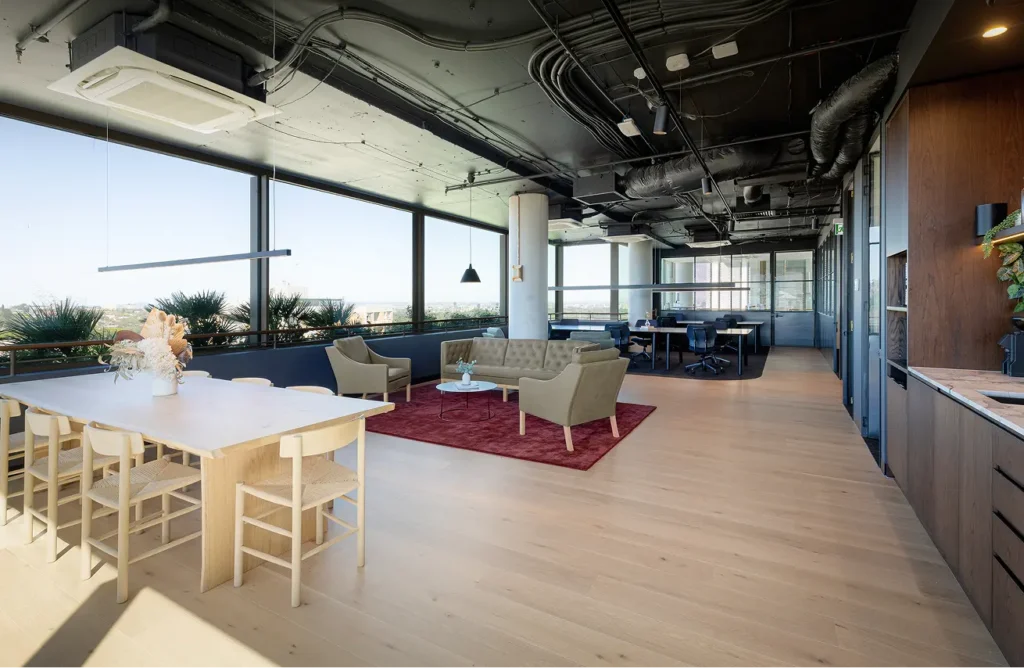 A modern open-plan office space features a dining table, lounge seating around a red rug, and work desks, surrounded by panoramic floor-to-ceiling windows and an industrial dark ceiling.