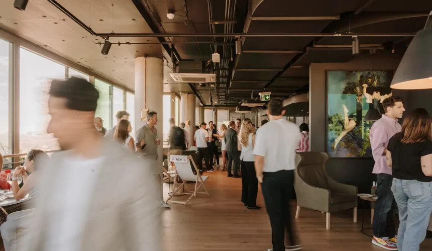 Well-dressed people socialize and mingle inside a modern, sunlit indoor lounge with large windows, dark industrial ceilings, and wooden flooring. Text on a white folding chair reads "AIX".