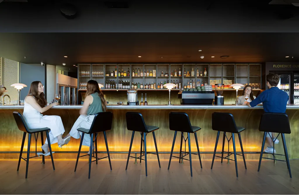 Four patrons sit on black stools, chatting and drinking along an illuminated brass bar counter backed by liquor shelves and espresso equipment. "FLORENCE GUILD" is written in the background.