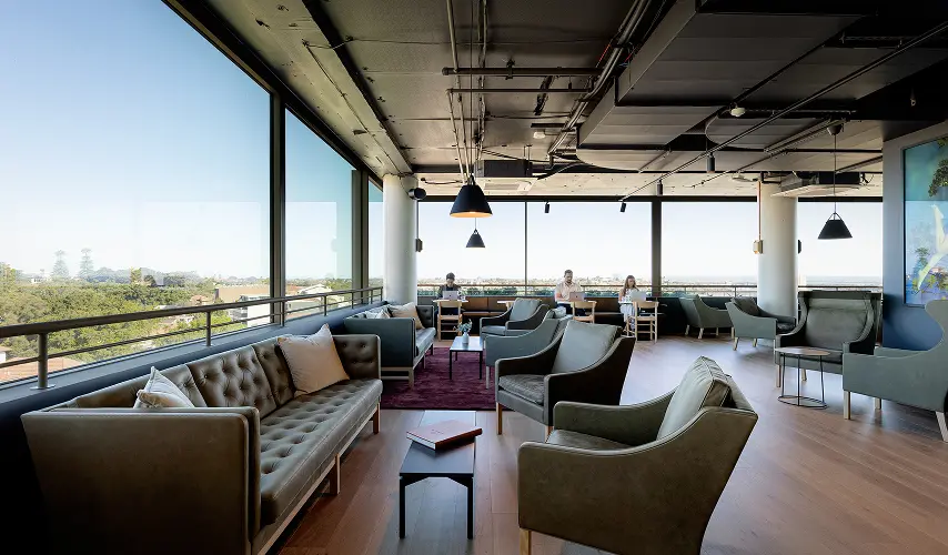 Leather sofas and armchairs furnish a spacious lounge where people work on laptops beside floor-to-ceiling windows overlooking a leafy green landscape under a clear sky.