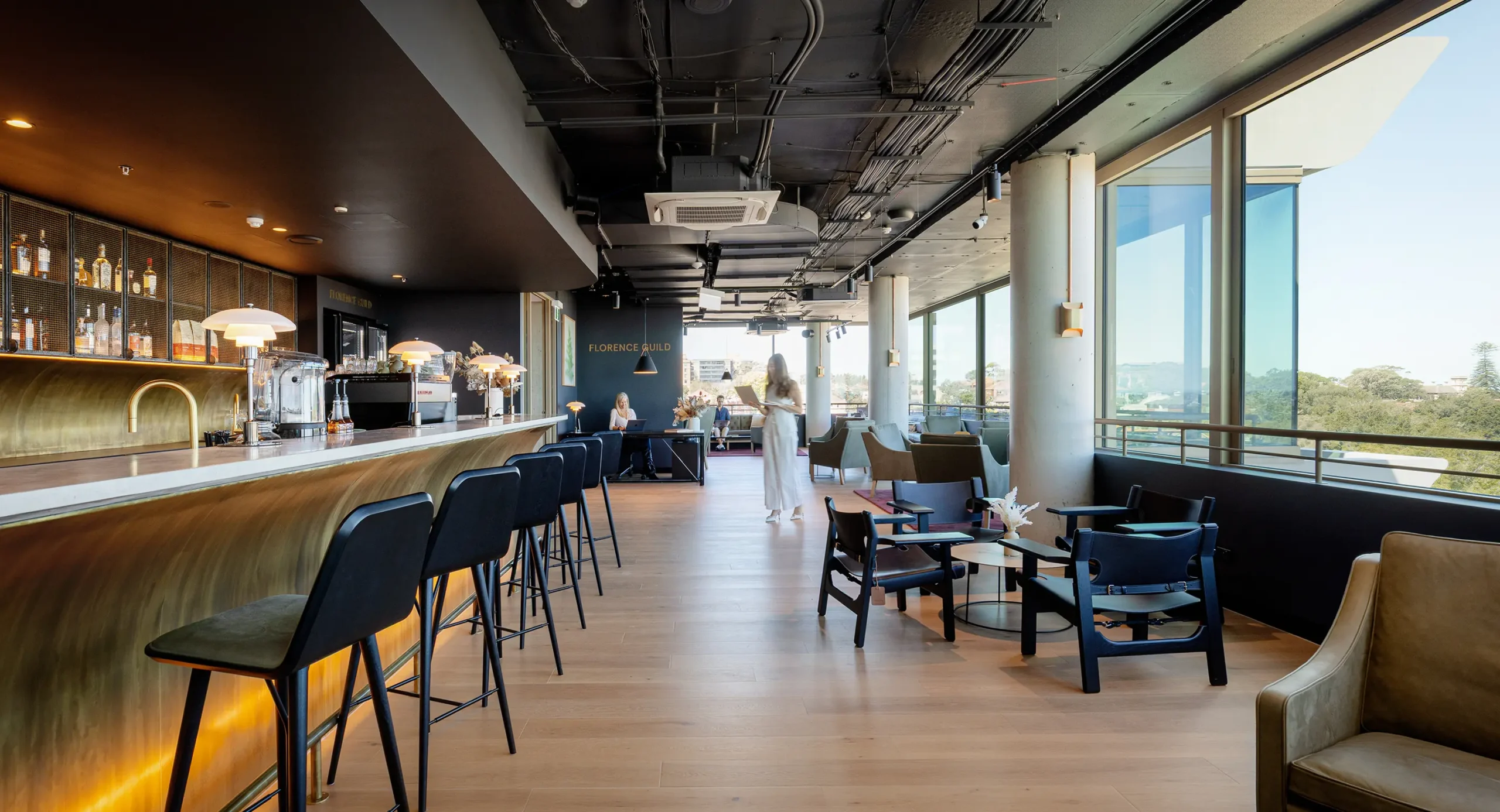 Patrons walk and work inside a modern lounge featuring a long brass bar with high stools, comfortable armchairs, exposed ceiling pipes, and expansive windows. Text in the image reads: "FLORENCE GUILD".