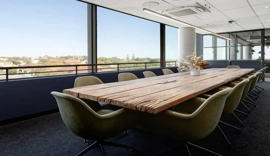 A long, rustic wooden conference table surrounded by olive-green swiveling armchairs stands inside a modern boardroom featuring floor-to-ceiling windows overlooking a sunny cityscape.