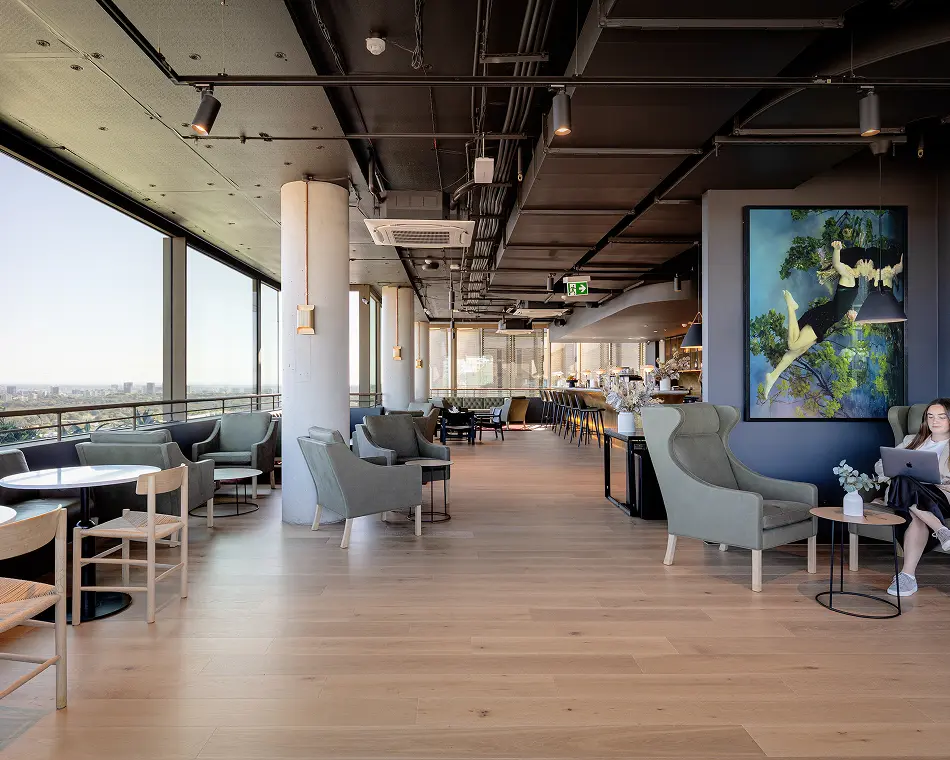 A woman sits using a laptop in a spacious, modern lounge filled with grey armchairs, light wood floors, and large panoramic windows overlooking a city skyline.