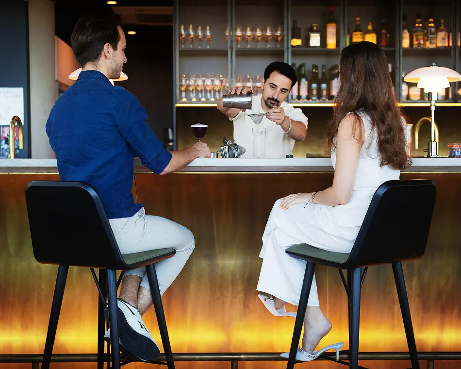 A bartender strains a cocktail behind a warm, illuminated bar counter, as a man in blue and a woman in white sit on high stools watching him prepare drinks.