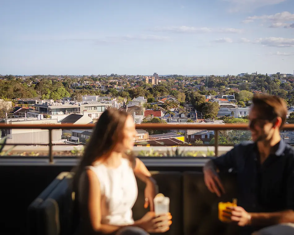 A couple sits blurred in the foreground on a rooftop lounge holding drinks, overlooking an expansive, sunny cityscape that stretches toward the horizon beneath a clear sky.