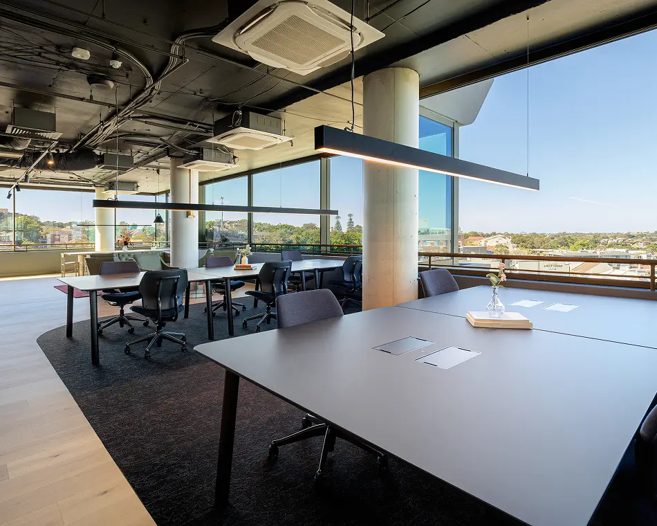 Modern grey office desks paired with black rolling chairs sit arranged in a spacious co-working room, featuring long linear pendant lights, dark carpets, and panoramic floor-to-ceiling city view windows.