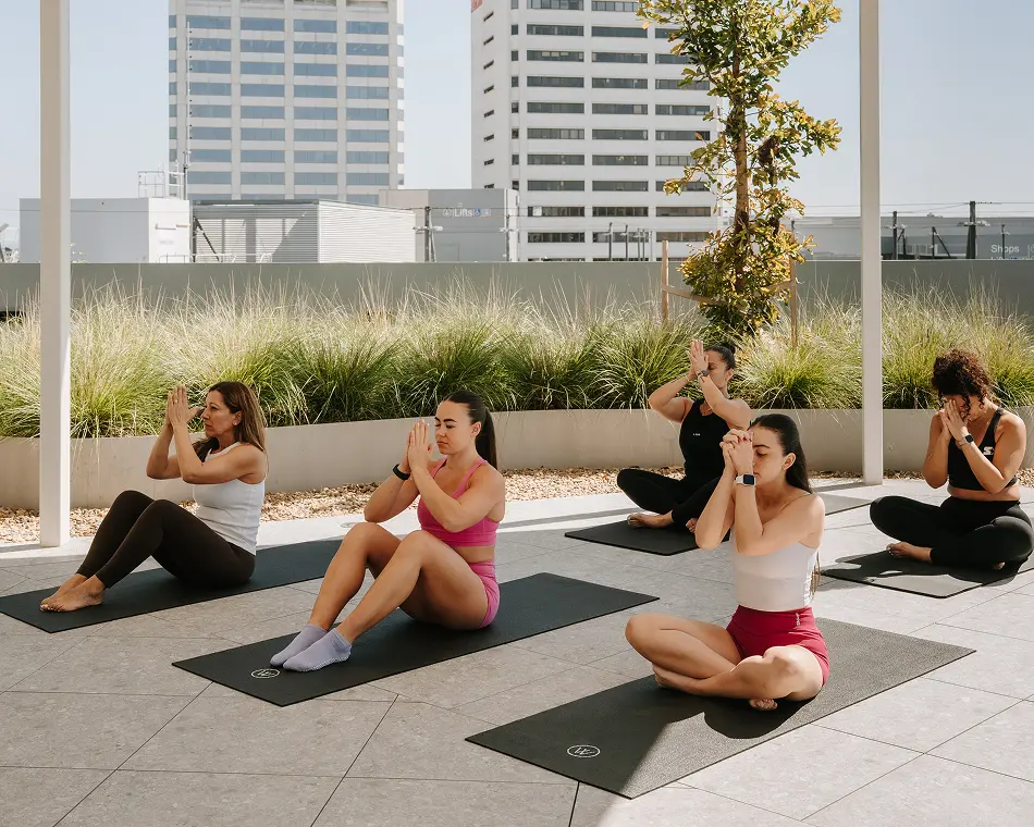 Five women in activewear practice yoga with hands pressed together on mats across a sunny rooftop terrace, set against greenery and city skyscrapers. Text in the background reads "Lifts" and "Shops".