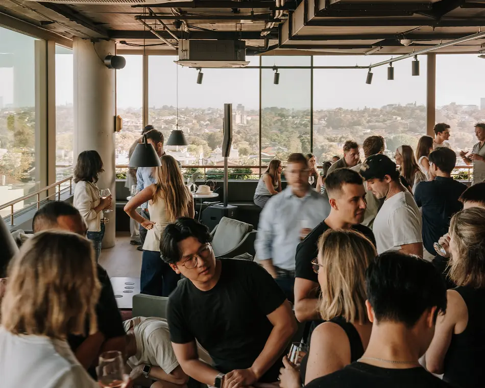 A crowd of people mingles and drinks inside a modern rooftop lounge featuring large floor-to-ceiling windows with panoramic views of a sunlit city landscape.