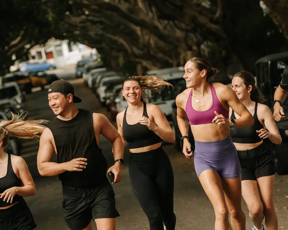 A group of smiling young adults in athletic wear runs together down a tree-canopied residential street lined with parked cars.