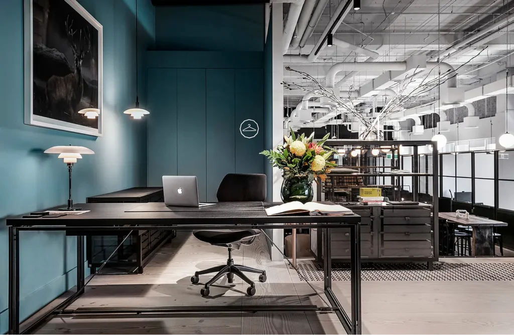 A dark desk equipped with an open laptop and chair stands in a modern workspace, bounded by teal walls and an exposed industrial ceiling. A yellow box reads, "TOOLKIT FOR LIFE".