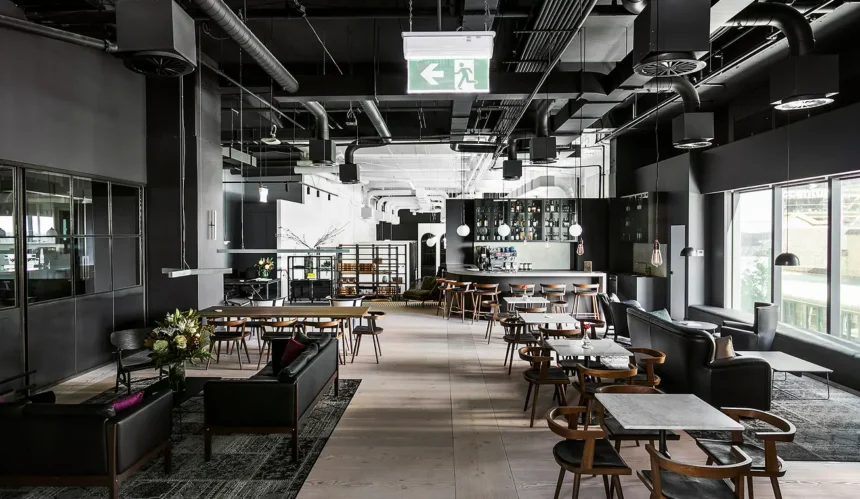 A modern interior lounge features black leather sofas, wooden dining sets, and a bar area, arranged across light wood flooring under an industrial black ceiling with exposed pipes.