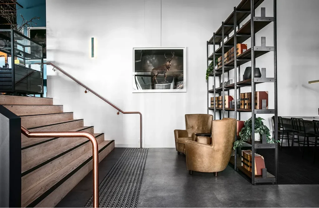 Wooden stairs with copper handrails ascend beside tan leather armchairs and a tall, filled metal bookcase within a modern interior featuring white walls and dark grey tile flooring.
