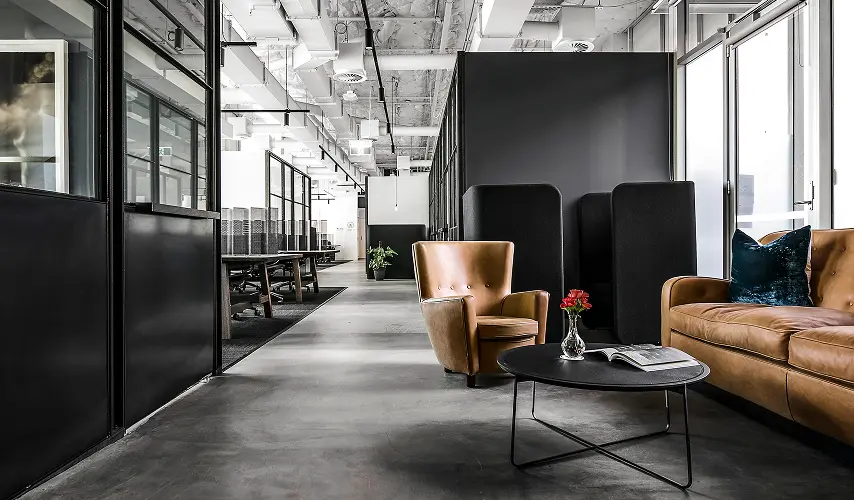 A tan leather armchair and sofa sit around a low coffee table inside a modern industrial office featuring exposed ceiling ducts, polished concrete floors, and dark glass partitions.