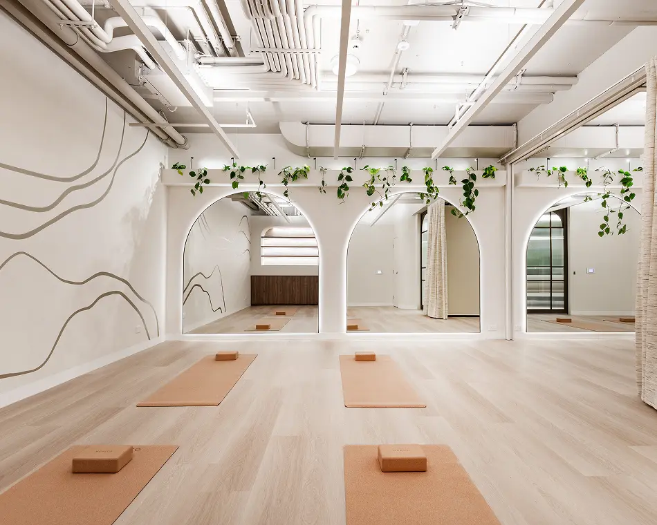 Tan yoga mats with cork blocks lay neatly arranged on a light wood floor inside a modern studio featuring arched mirrors, hanging greenery, and exposed white ceiling pipes.