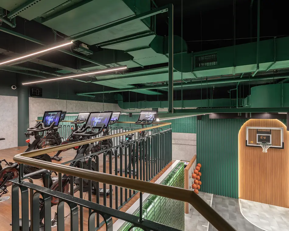 Stationary exercise bikes are lined up on a wooden mezzanine floor within a modern gym featuring exposed green ceiling pipes, a metal railing, and a basketball hoop on the lower level.