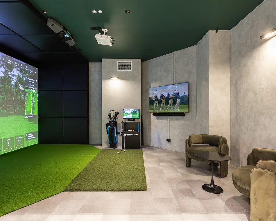 An indoor golf simulator projects a virtual golf course onto a large screen, positioned beside artificial turf mats, golf clubs, and velvet armchairs within a sleek modern room. Text reads: "FORESIGHT SPORTS".