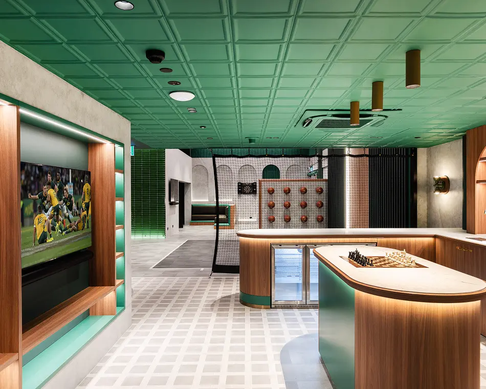 A chessboard sits on a wooden island inside a modern entertainment room, positioned near a wall-mounted TV showing rugby, beneath a bold green coffered ceiling.
