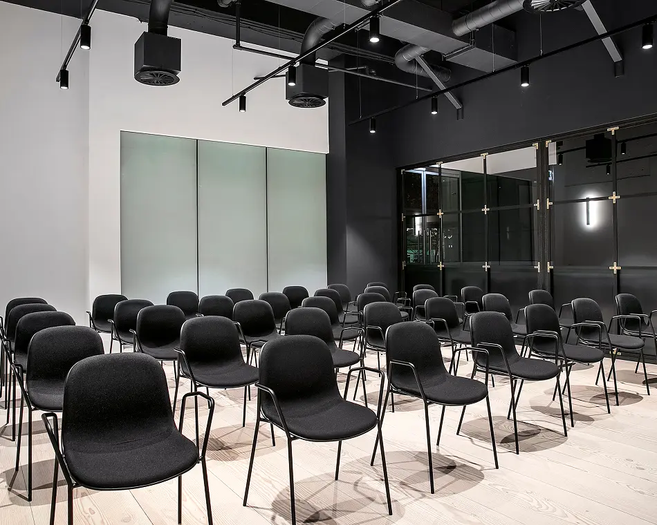 Rows of sleek black chairs face forward in a modern presentation room featuring light hardwood floors, contrasting black and white walls, track lighting, and exposed ceiling ductwork.