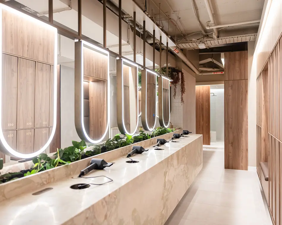 Black hair dryers sit ready for use along a marble vanity lined with green plants beneath illuminated U-shaped mirrors, inside a modern wood-paneled locker room featuring exposed ceiling pipes. Reflected locker doors display numbers 020, 021, 022, 023, 024, and 025, while a distant red sign reads "FIRE HYDRANT".