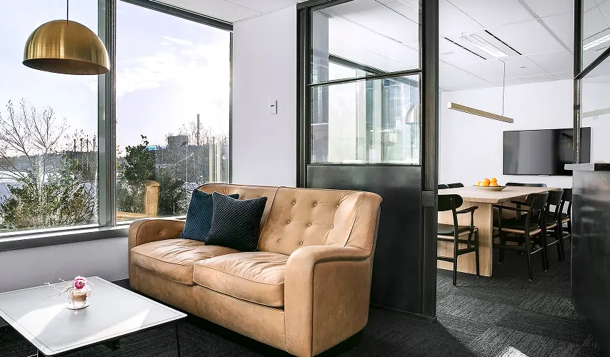 A tan leather sofa adorned with dark blue pillows sits in a modern office lounge beside floor-to-ceiling windows, next to a glass-partitioned meeting room with a wooden table.