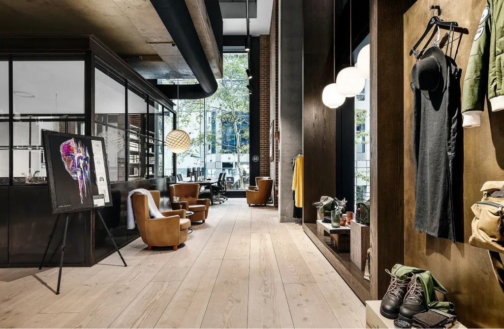 Brown leather armchairs, an easel displaying a colorful portrait, and boutique clothing displays line a light-wood corridor inside a modern industrial space with glass partitions and tall windows.
