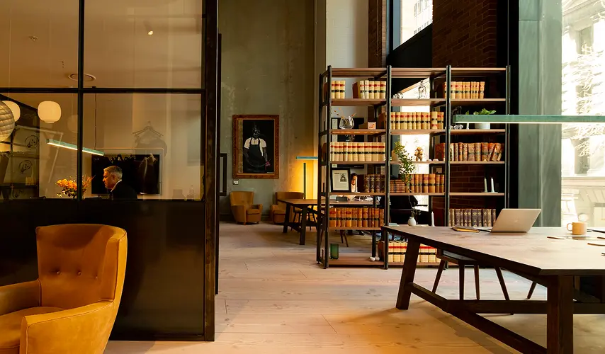 A yellow leather armchair and wooden worktables sit in a modern, open-plan office interior featuring tall metal bookshelves, glass partitions, high ceilings, and warm ambient lighting.
