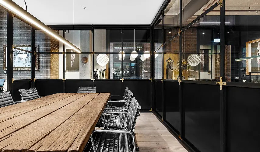 A long wooden conference table surrounded by black leather office chairs sits inside a modern meeting room enclosed by glass wall partitions with overhead linear lighting.