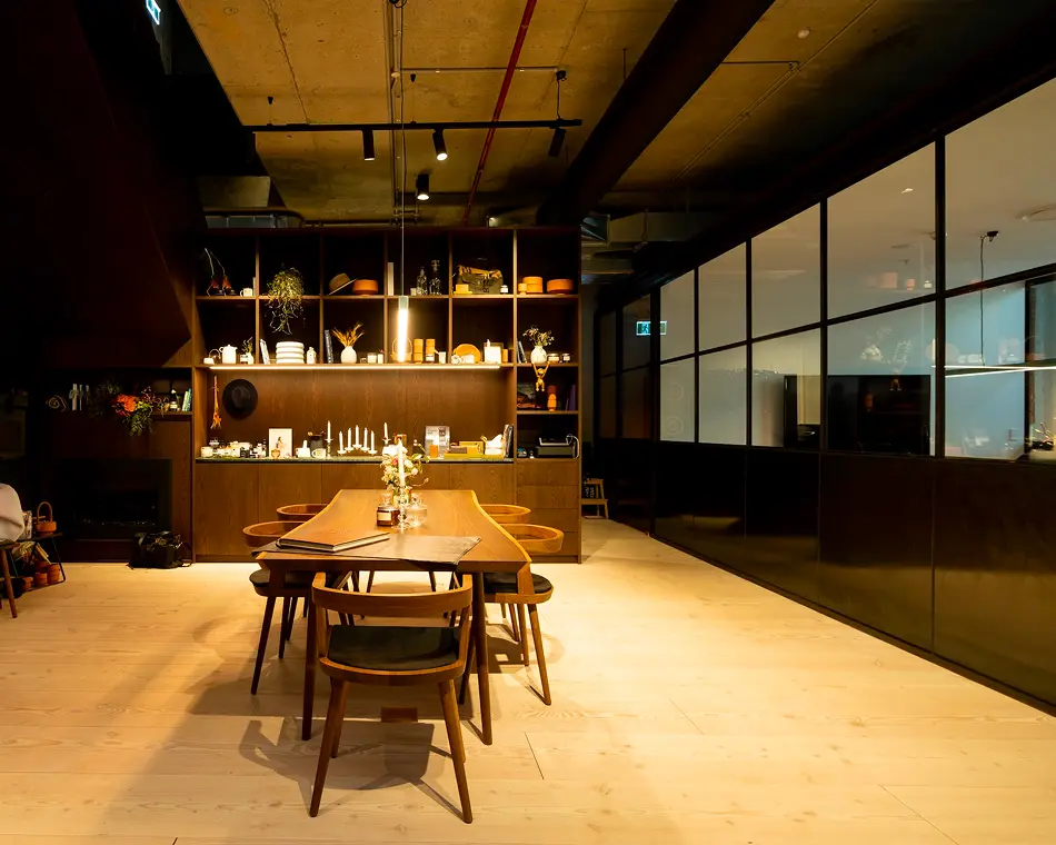 A long wooden dining table surrounded by chairs sits illuminated by warm lighting inside a modern room featuring light wood floors, dark shelving filled with decor, and a glass partition.