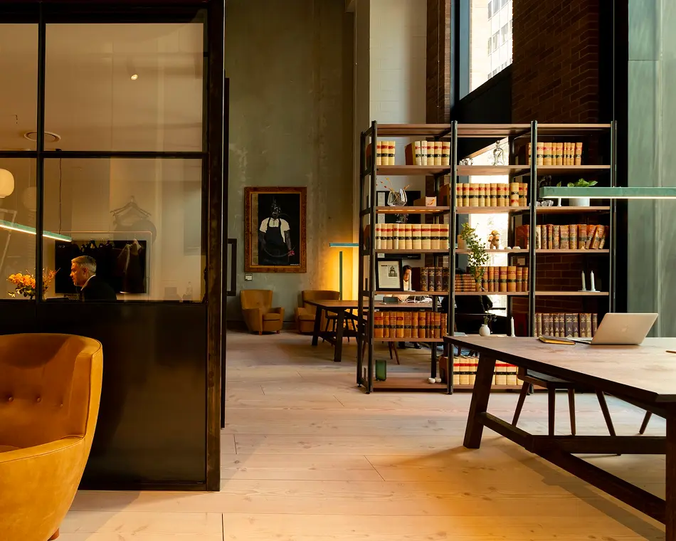 Tall shelving units filled with bound books stand beside wooden study tables within a spacious, industrial-style lounge featuring glass-partitioned rooms, leather armchairs, high ceilings, and light wood flooring.
