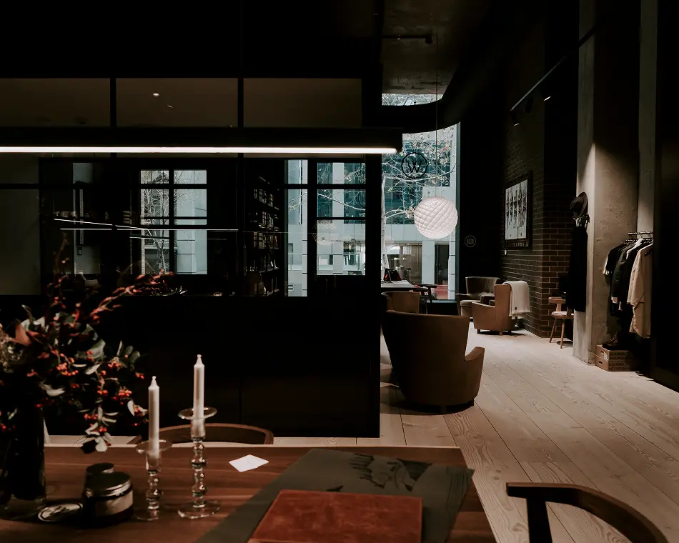 A dark wooden table adorned with white taper candles and floral arrangements sits illuminated beneath a linear pendant light within a dim, upscale lounge featuring armchairs and light wood floors.