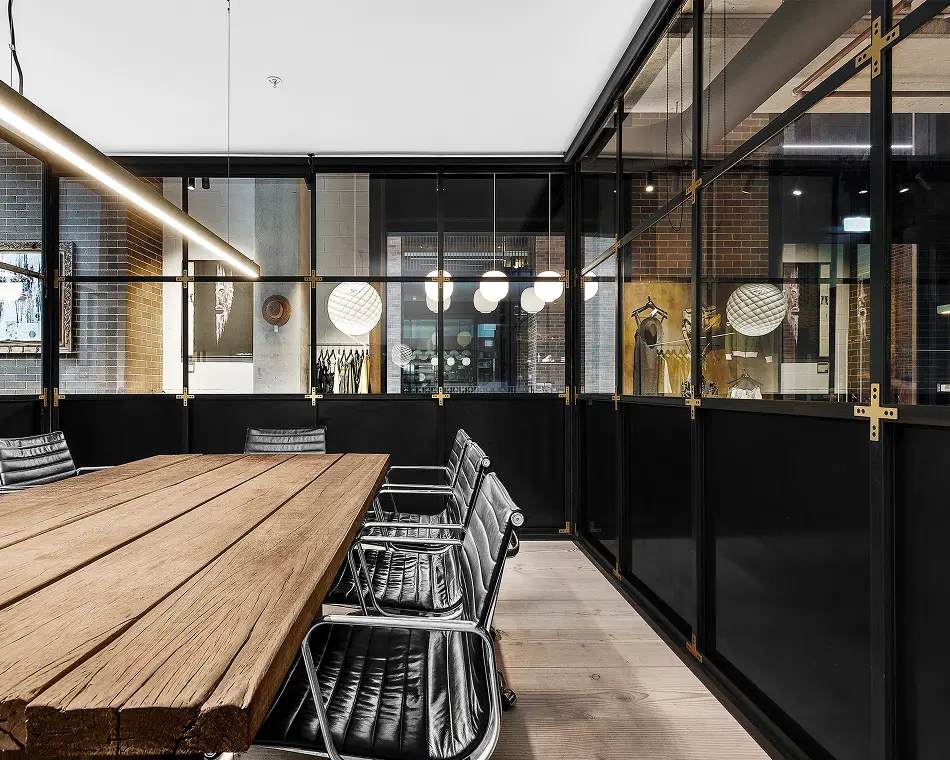 A long, rustic wooden conference table surrounded by black leather office chairs sits inside a modern meeting room enclosed by industrial black-framed glass partitions with brass accents.