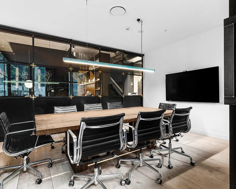 A long wooden conference table surrounded by black leather executive chairs stands inside a modern meeting room featuring glass partition walls, a wall-mounted TV, and a linear pendant light.