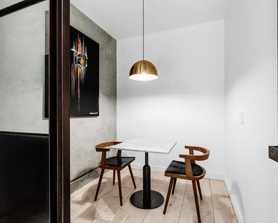 A marble-topped table with two wooden chairs sits beneath a brass dome pendant light inside a modern room featuring light wood floors and a concrete wall with abstract art.