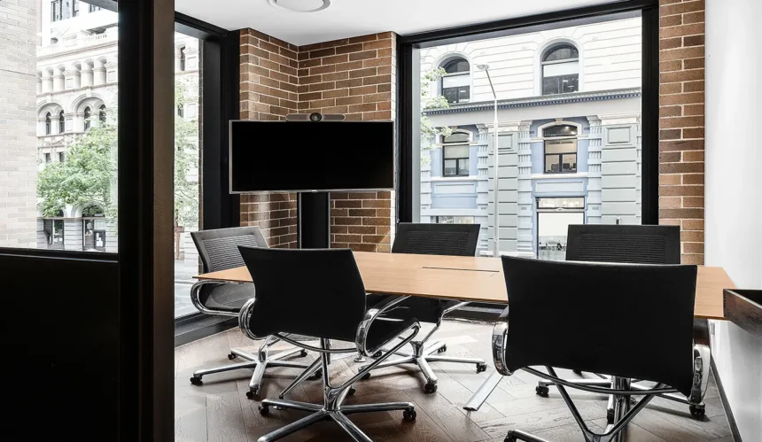 A wooden conference table surrounded by black rolling chairs stands in a modern meeting room with exposed brick walls, a TV monitor, and large windows overlooking city buildings.