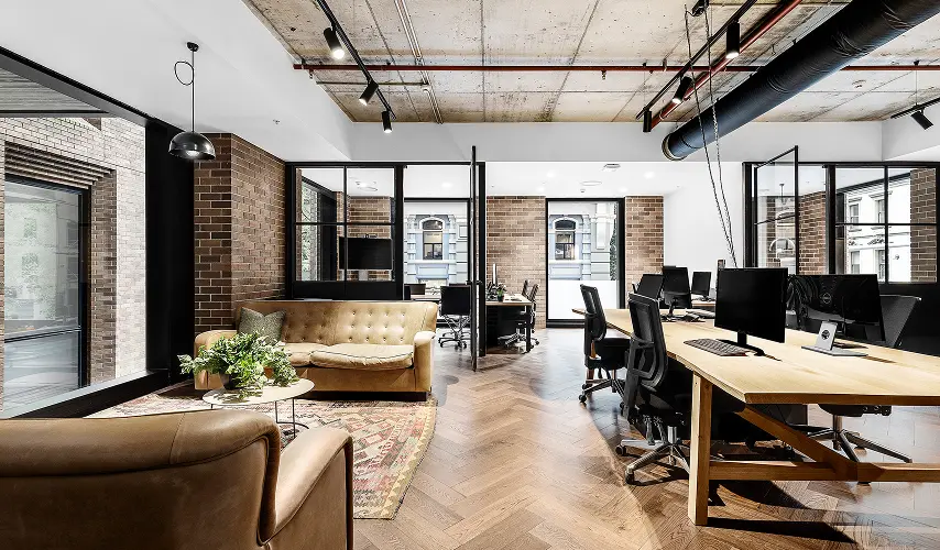 A modern industrial office features a lounge area with leather sofas beside a long wooden desk with computer workstations, set on herringbone floors beneath exposed brick and concrete ceilings.